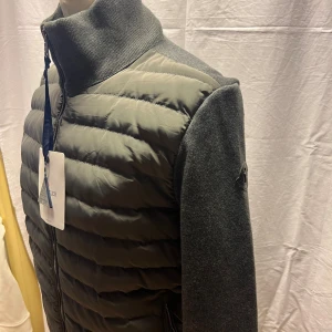Grå pufferjacka från Moncler - Snygg grå pufferjacka från Moncler med quiltad framsida och stickade ärmar och rygg. Jackan har hög krage, dragkedja framtill och Moncler-logga på ärmen. Perfekt för dig som vill ha en stilren och modern look.