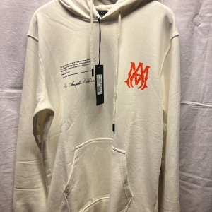 Vit hoodie från Amiri med tryck - Snygg vit hoodie från Amiri med svart och orange tryck både fram och bak. Tröjan har huva med snörning, känguruficka och coola grafiska detaljer. Tillverkad i 100% bomull för en skön och avslappnad känsla.