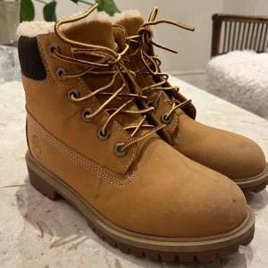 Timberland boots med ullfoder - Ny pris: 2426kr. Klassiska Timberland boots i ljusbrunt skinn med grov sula och snörning. Det är mycket sparsamt andvända och Skorna har mörkbrunt parti vid hälen och är fodrade med mjuk ull för extra värme. Perfekta för kalla dagar och har en robust, ikonisk look.