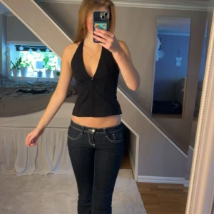 Svart halterneck korsett-topp - Säljer en svart korsett-topp med halterneck och djup v-ringning. Toppen är croppad och har en snygg passform som framhäver midjan. Stängs med ”knappar” framtill och har breda band. Det är sytt i nacken så att banden blir kortare men går nog att sprätta bort. Perfekt för dig som vill ha en stilren och trendig look.