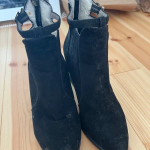 Svarta mocka boots med klack - Snygga svarta boots i mocka med markerad klack och spetsig tå. Skorna har dragkedja bak och ett dekorativt spänne runt ankeln. Perfekta för dig som vill ha en stilren och trendig look.