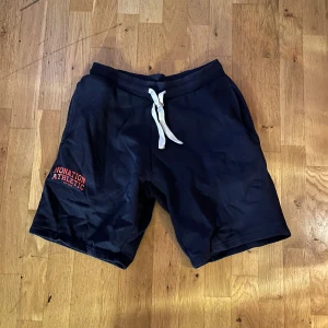 Svarta shorts Nonation Athletic - Svarta shorts från Nonation Athletic med vit snörning i midjan och röd logga på vänster ben. Mjukt material och avslappnad passform, perfekta för träning eller chill. Två sidofickor och elastisk midja för extra komfort.