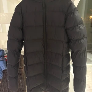 Svart lång dunjacka från Neomondo - Säljer en svart dunjacka från Neomondo med huva och dragkedja framtill. Jackan är lång i modellen och har puffiga, quiltade sektioner som ger en schysst look. Perfekt för kalla dagar och enkel att matcha med jeans eller mjukisar.