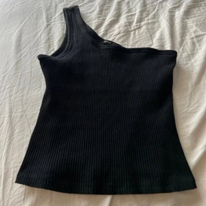 Svart ribbad oneshoulder topp - Snygg svart ribbad topp med oneshoulder-design. Toppen är ärmlös och har en tight passform som framhäver siluetten. Perfekt för att skapa en trendig look med enkel twist. Materialet är mjukt och stretchigt för extra komfort.