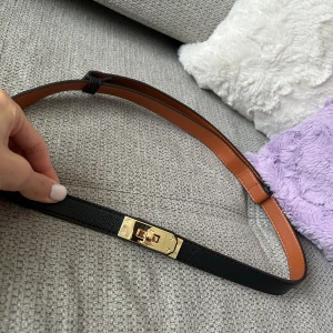 Svart smalt skärp med gulddetalj - Snyggt och stilrent svart bälte i skinn med smal design och exklusivt guldfärgat spänne framtill. Insidan är brun och bältet har en elegant struktur på ytan. Perfekt accessoar för att lyfta din outfit med en lyxig touch.