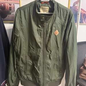 Olivgrön bomberjacka Jack & Jones - Snygg olivgrön bomberjacka från Jack & Jones med dragkedja framtill, ribbade muddar och två fickor med dragkedja. Jackan har en klassisk bomberkrage och en liten patch på bröstet. Perfekt för dig som gillar en clean och stilren look.