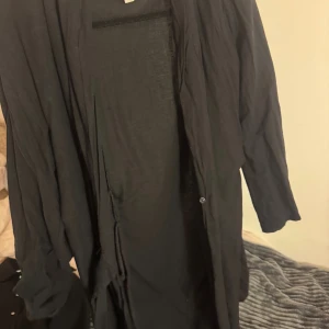 Svart lång overshirt  - Säljer en svart lång overshirt med knappar framtill. Jackan har en avslappnad passform och är tillverkad i ett tunt, mjukt material. Perfekt att slänga över en t-shirt för en chill look.