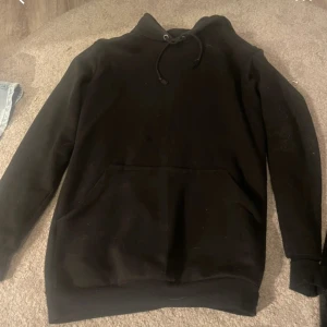 Svart hoodie med huva och ficka - Svart hoodie med klassisk huva och snörning, samt stor magficka framtill. Tröjan har långärmade ärmar och är gjord i mjukt material som passar perfekt för chill dagar. Enkel och stilren design som funkar till det mesta.