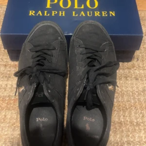 Svarta sneakers från Polo Ralph Lauren - Svarta sneakers från Polo Ralph Lauren med klassisk snörning och rund tå. Skorna är tillverkade i canvas med Polo-logga broderad på sidan och brun gummisula med Polo-tryck undertill. Perfekta för dig som gillar stilren och sportig look.