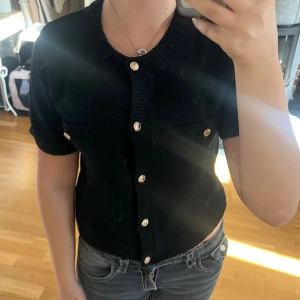 Svart stickad blus med guldfärgade knappar - Säljer en svart stickad blus med korta ärmar och rund hals. Toppen har snygga guldfärgade knappar framtill och på de två bröstfickorna. Modellen är croppad och har ribbade detaljer vid hals, ärmslut och nederkant. Perfekt för dig som gillar klassisk stil med en twist.