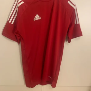 Röd Adidas fotbollströja Aeroready - Röd fotbollströja från Adidas med klassiska vita ränder på axlarna och Adidas-logga på bröstet. Tillverkad i lätt och ventilerande Aeroready-material som passar perfekt för träning eller match. Kortärmad modell med rund hals.