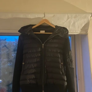 Moncler cardigan  - Svart pufferjacka från Moncler med stickade ärmar och huva. Jackan har quiltad front, dragkedja hela vägen och en ficka med dragkedja på ärmen. Klassisk Moncler-logga på ena ärmen. Perfekt för kyliga dagar och snygg till streetwear.
