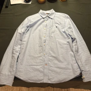 Ljusblå skjorta från Morris S - Säljer en stilren ljusblå skjorta från Morris i storlek S. Skjortan har klassisk krage, knappar framtill och broderad logga på bröstet. Tillverkad i mjuk bomull, perfekt för en clean och snygg look.