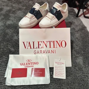 Valentino Garavani Open sneakers vit/blå - Säljer ett par Valentino Garavani Open sneakers i vitt skinn med bred marinblå rem över vristen och ikoniska nitar på hälen. Klassisk rund tå, platt sula och snörning framtill. Märkeslogga på plös och sula. Kommer med originalpåse och certifikat.
