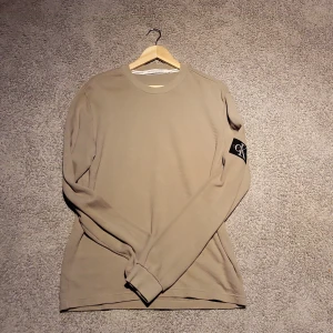Beige långärmad tröja Calvin Klein - Stilren beige långärmad tröja från Calvin Klein Jeans med svart CK-logga på ärmen. Tröjan har rund halsringning och är tillverkad i ett mjukt, ribbat bomullsmaterial. Perfekt för dig som gillar enkel och clean look.