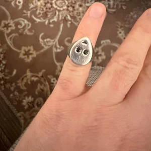 Maria Nilsdotter 14.25mm - Unik silverring med droppformad topp och ett ingraverat smiley-ansikte. Ringen har två svarta stenar som ögon och en liten böjd mun, vilket ger den en cool och lekfull vibe. Perfekt accessoar för dig som gillar statement-smycken och vill sticka ut.