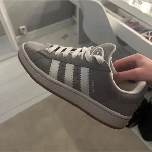 Grå adidas Campus sneakers  - Säljer ett par grå adidas Campus sneakers med klassiska vita ränder på sidorna och vit snörning. Skorna har en rund tå och platt sula, tillverkade i mocka med gummisula. Perfekt för dig som gillar stilrena och tidlösa sneakers.