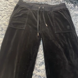 Svarta mjukisbyxor Juicy Couture M - Svarta mjukisbyxor från Juicy Couture i storlek M. Byxorna har bred resår i midjan med snörning, och en diskret Juicy Couture-logga på ena fickan. Mjukt och glansigt sammetstyg som känns lyxigt och skönt.