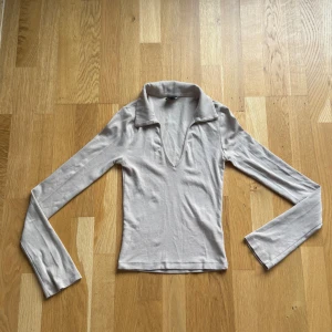 Snygg beige långärmad topp från Gina Tricot - Säljer en beige långärmad topp från Gina Tricot i mjuk bomull med lite stretch. Toppen har en snygg krage och djup v-ringning framtill, vilket ger en cool och trendig look. Perfekt att styla med jeans eller kjol för en chill vibe.