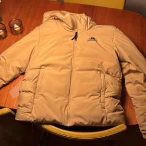 Beige pufferjacka från Adidas - Säljer en beige pufferjacka från Adidas med huva och svarta loggan på bröstet samt tre svarta ränder på ryggen. Jackan har dragkedja framtill och två stora fickor. Perfekt för kyliga dagar och har en clean streetstyle-look. Den har några svarta små fläckar men annars dunder skick.