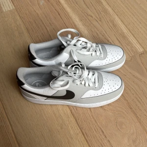 Nike Court Vision Low sneakers vit/svart - Nike Court Vision Low sneakers i vit och grått med svarta detaljer och klassisk swoosh på sidan. Skorna har snörning, perforerad tå och platt sula. Tillverkade i syntet och skinn för en clean och sportig look. Perfekta för dig som gillar stilrena sneakers.