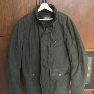 Mörkgrön quiltad jacka Oscar Jacobson - Säljer en mörkgrön jacka från Oscar Jacobson i vaxad bomull med quiltat foder. Jackan har dragkedja, tryckknappar och flera fickor framtill. Klassisk modell med hög krage och snygga detaljer. Perfekt för dig som gillar stilren och funktionell design.