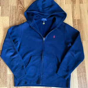 Marinblå hoodie från Polo Ralph Lauren - Säljer en marinblå zip-hoodie från polo Ralph lauren. Passar 165 till 175cm. Tröjan är i topp skick då den endast används en fåtal gånger. Tröjan har inga tecken på användning. Säljer på grund av att jag har växt ur den. Köpt för ca 1000kr. 