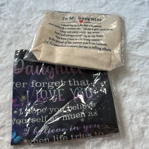 Necessär och shoppingväska med text - Säljer ett set med en beige necessär med tryckt text 'To My Daughter' och hjärtan, samt en svart shoppingväska med färgglada fjärilar och inspirerande text i lila och blått. Perfekt som present till dottern, båda med unika motiv och budskap.