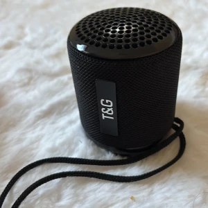 T&G Mini Bluetooth Högtalare - Kompakt och portabel T&G Bluetooth-högtalare i svart. Perfekt för resor eller utomhusbruk med praktisk handledsrem. Högtalaren är i mycket gott skick utan synliga repor eller skador. Enkel att använda med tydliga knappar på undersidan.