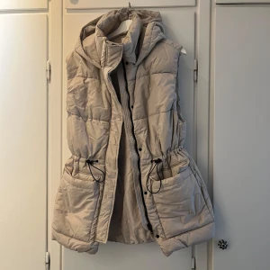 Beige  dunväst med huva - Dunväst från HM med stor huva och snörning i midjan. Västen har två  fickor framtill samt på sidan och stängs med dragkedja och knappar. 