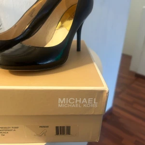 Svarta pumps från Michael Kors - Snygga svarta pumps från Michael Kors med rund tå och hög klack. Skorna är tillverkade i blankt syntetmaterial med guldfärgad innersula och en liten gulddetalj bak på hälen. Perfekta för dig som vill ha en elegant och stilren look.