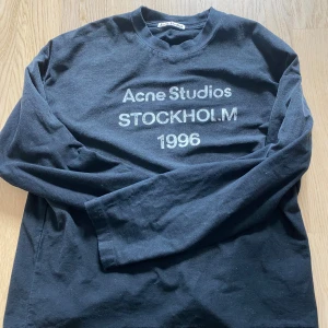 Svart Acne Studios långärmad tröja  - Svart långärmad tröja från Acne Studios med vit text 'Acne Studios STOCKHOLM 1996' på bröstet. Rund hals och relaxed passform, tillverkad i mjuk bomull. Perfekt för dig som gillar stilrena och klassiska streetwear-plagg.