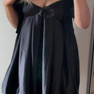 Svart kort satinklänning med v-ringning - Supersnygg svart kort klänning i satin med v-ringad byst och lätt utställd passform. Klänningen har korta ärmar och en söt rosettdetalj framtill. Perfekt för dig som gillar en stilren och chic look.
