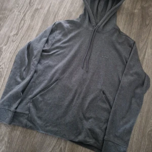 Mörkgrå hoodie från BOSS - Snygg mörkgrå hoodie från BOSS med diskret logga på bröstet. Tröjan har huva med snörning, känguruficka och långa ärmar. Perfekt för chill dagar och enkel att matcha med jeans eller joggers. Märket är reflex