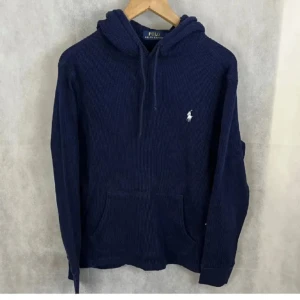 Marinblå stickad hoodie Polo Ralph Lauren - Snygg marinblå stickad hoodie från Polo Ralph Lauren med klassisk huva och snörning. Tröjan har en broderad vit logga på bröstet och en stor magficka framtill. Perfekt för dig som gillar stilren och bekväm streetwear.