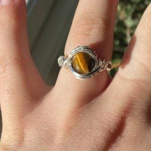 Silvrig ring med tigeröga-sten - Unik handgjord ring med en rund tigeröga-sten i centrum, infattad i snirkligt silverfärgat wire. Stenen har en snygg brun och gul ton som skiftar i ljuset. Perfekt för dig som gillar boho-vibe och naturinspirerade smycken.
