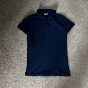 Blå merino pikétröja från John Henric - Snygg marinblå pikétröja från John Henric med klassisk krage och tre knappar framtill. Tröjan har korta ärmar och är tillverkad i mjukt material som känns skönt mot huden. Perfekt för en stilren och avslappnad look.