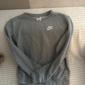 Grå sweatshirt från Nike XS - Snygg grå sweatshirt från Nike i storlek XS. Tröjan har rund hals, långärmade ärmar och en vit Nike-logga broderad på bröstet. Mjuk insida och klassisk passform, perfekt för chill eller träning. Materialet är bomull och polyester.