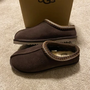 Bruna UGG Tasman slip-ins i mocka - Mysiga bruna slip-ins från UGG i mjuk mocka med fluffigt fårskinnsfoder. Skorna har dekorativt mönster runt öppningen och en rejäl sula som funkar både inne och ute. Perfekta för kalla dagar när du vill ha det extra bekvämt. Mindre i storlek passar därför bättre som 43 än 44.