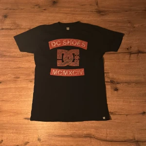 Svart DC Shoes t-shirt med röd logga - Svart t-shirt från DC Shoes med stor röd logga och texten 'DC SHOES' samt 'MCMXCIV' på bröstet. Klassisk passform och rund hals. Perfekt för dig som gillar streetwear och skatekänsla.