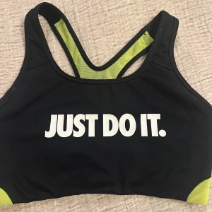 Nike svart och limegrön sporttopp - Nike Dri-Fit sporttopp i svart med limegröna meshdetaljer och vit 'JUST DO IT.' print framtill. Swoosh-logga på ryggen. Tight passform och ärmlös design, perfekt för träning. Materialet är syntet med fuktavvisande egenskaper för extra komfort.