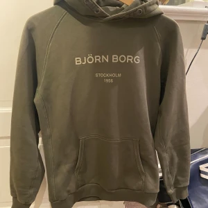 Olivgrön hoodie från Björn Borg - Snygg olivgrön hoodie från Björn Borg med tryck på bröstet. Tröjan har huva med snörning och en stor magficka framtill. Mjuk insida och klassisk passform, perfekt för chill eller skola.
