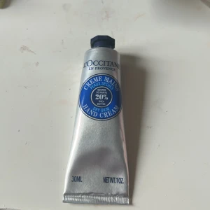 L'Occitane handkräm med sheasmör - Handkräm från L'Occitane med 20% sheasmör, perfekt för torra händer. Tuben är silverfärgad med blå etikett och rymmer 30 ml. Krämen har en mjuk konsistens och kommer i en praktisk metallförpackning med skruvlock.