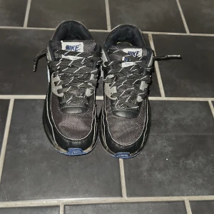 Nike Air Max 90 svart/blå sneakers - Säljer ett par Nike Air Max 90 sneakers i svart med blå och grå detaljer samt vit swoosh. Skorna har snörning, platt sula och är tillverkade i en mix av syntet och mesh för en sportig look. Perfekta för dig som vill ha en fet drip.