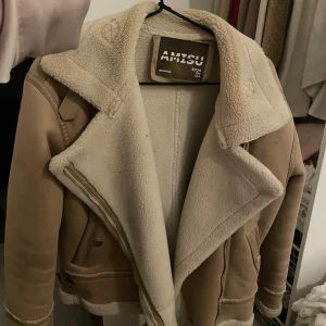 Beige teddyjacka från Amisu - Säljer en beige teddyjacka från Amisu med fluffigt foder och bred krage. Jackan har snygga detaljer som dragkedja, knappar och bälte vid nederkanten. Perfekt för dig som vill ha en varm och trendig look i höst och vinter.