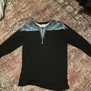 Svart tröja med blått vingtryck Marcelo Burlon - Svart långärmad tröja från Marcelo Burlon County of Milan med ett fett blått och vitt vingtryck över axlar och hals. Tröjan har rund hals och är gjord i mjuk bomull. Perfekt för dig som vill sticka ut med en unik design.