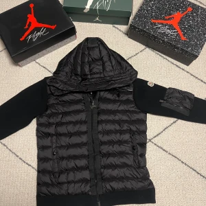 Moncler Cardigan - Helt ny aldrig använd svart pufferjacka från Moncler med stickade ärmar och vadderad kropp. Jackan har huva, dragkedja framtill och en ficka med dragkedja på ena ärmen. Klassisk Moncler-logga på ärmen och ribbade muddar. Perfekt för dig som vill ha en stilren och sportig look. Denna kan verkligen aldrig gå fel oavsett vilken stil du har.
