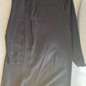Svart stickad långärmad klänning XS - Svart stickad klänning i storlek XS med lång ärm och ribbade muddar vid hals och ärmslut. Klänningen har en enkel, rak siluett och en snygg detalj med liten rynkning i midjan. Perfekt för dig som gillar stilrena och bekväma plagg.