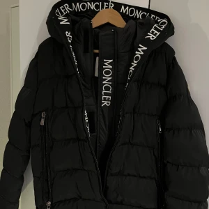 Svart Moncler dunjacka med huva - Svart pufferjacka från Moncler med stor huva och tydlig logotyp på både insidan och ärmen. Jackan är ny och aldrig använd med prislapp på. Jackan har dragkedjor på insidan och utsidan samt coola detaljer som tryckt Moncler text på huvan och längs dragkedjan. Pris går att diskutera!🤗