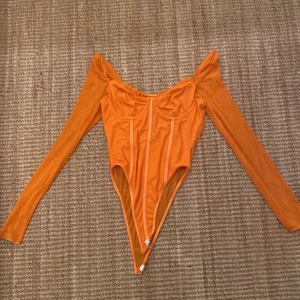 Orange långärmad korsettbody - Säljer en orange långärmad korsettbody med offshoulder-design och markerade sömmar framtill. Bodyn har kupor med lätt rynk upptill och knäppning i grenen. Materialet är tunt och stretchigt, perfekt för att skapa en snygg siluett.